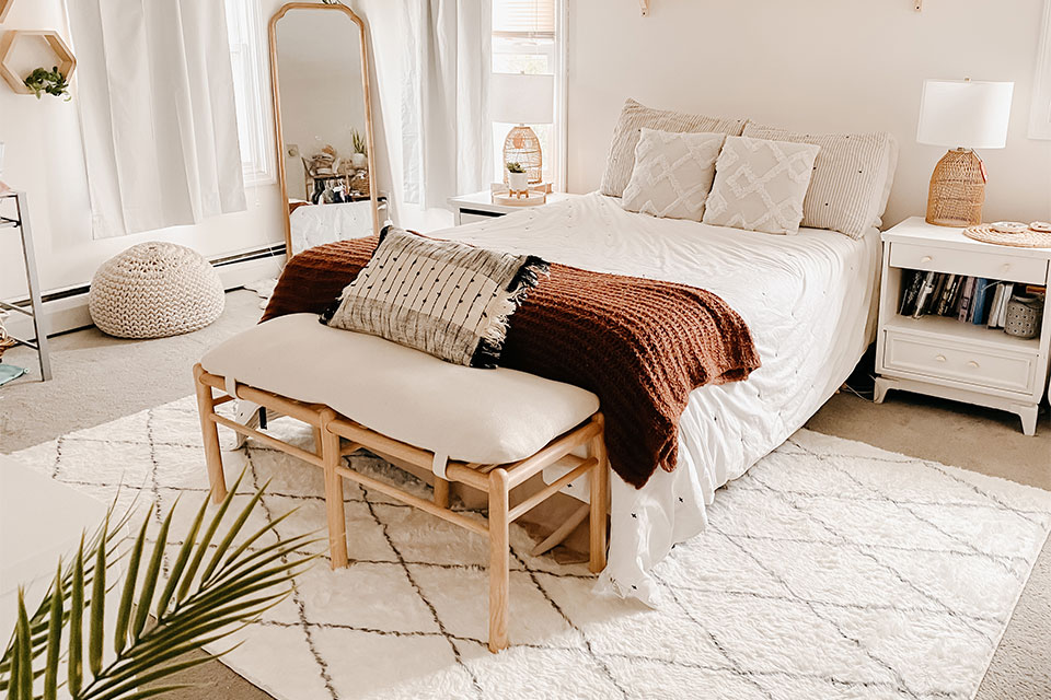 White plush rug with diamond pattern under a white queen bed with plants and wooden bench
