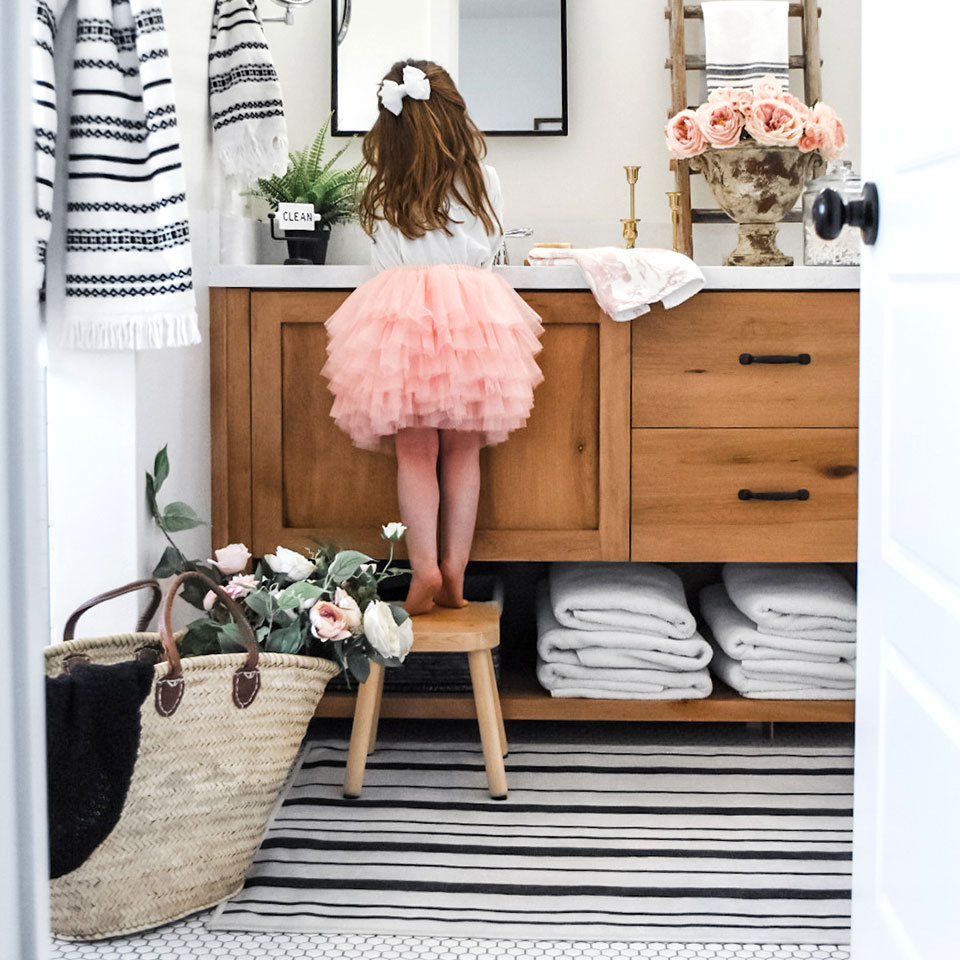 Girl in pink skirt by bathroom counter with flowers and black and white striped rug