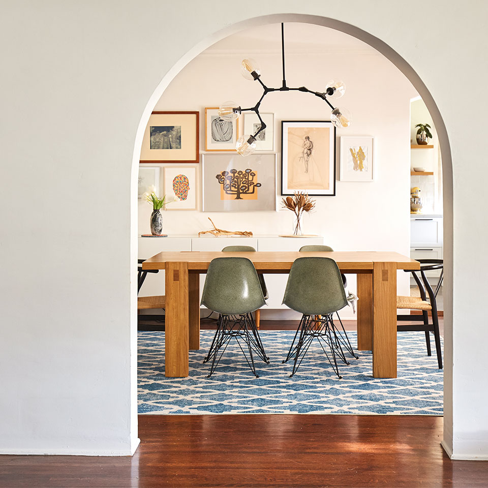 Wooden dining table with green midcentury modern chairs on blue diamond rug with artworks on white wall.