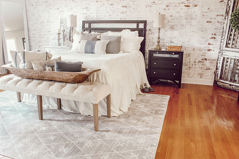 Grey diamond rug with white bed cream bench and brick wall in farmhouse bedroom