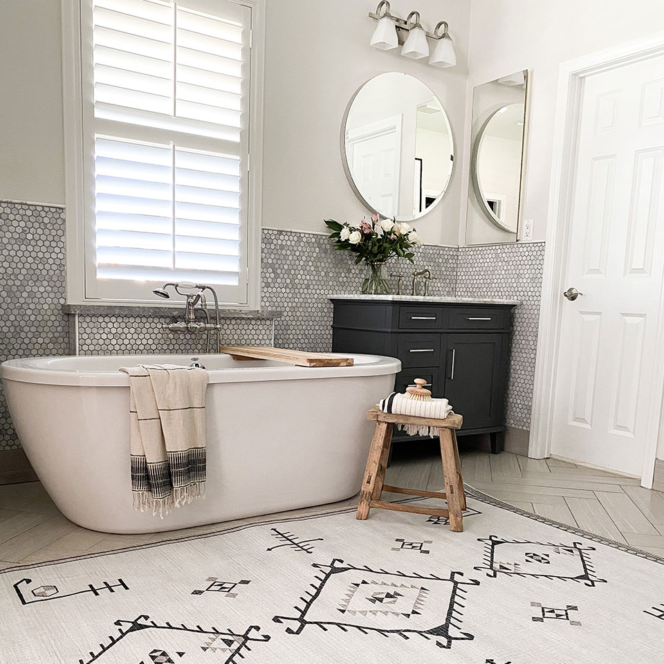 Black and white geometruc rug by cream bathtub with wooden stool and round mirror