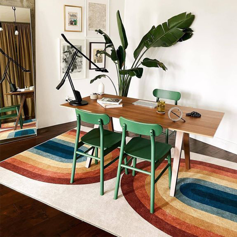 Rainbow rug under wood table with green chairs plant and mirror.