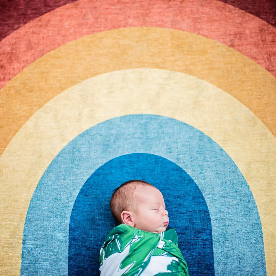Rainbow rug with newborn baby in green swaddle.