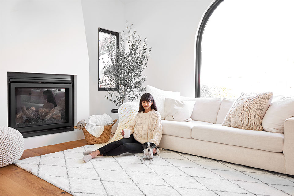 Girl with white sweater in a white living room with white couch white rug white walls and puppy by the fireplace